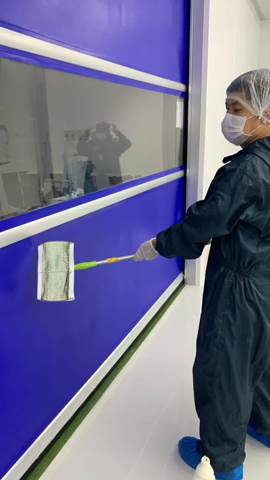 Technician wipe-cleaning a cleanroom door in full PPE